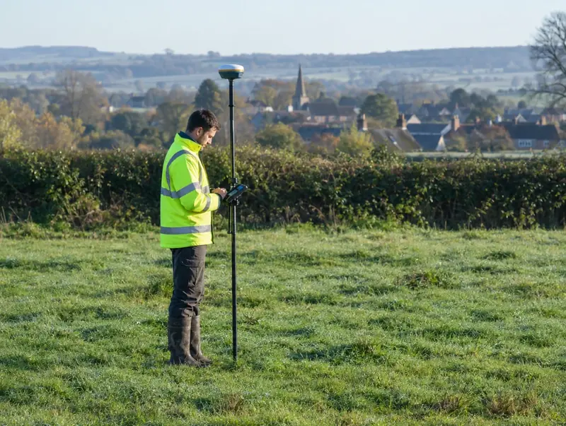 Surveyor in hi-vis using a GPS rover pole in a grass field near a village